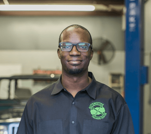 Certified auto technician Larry Witherspoon Jr. stands in an auto body shop with a car behind him