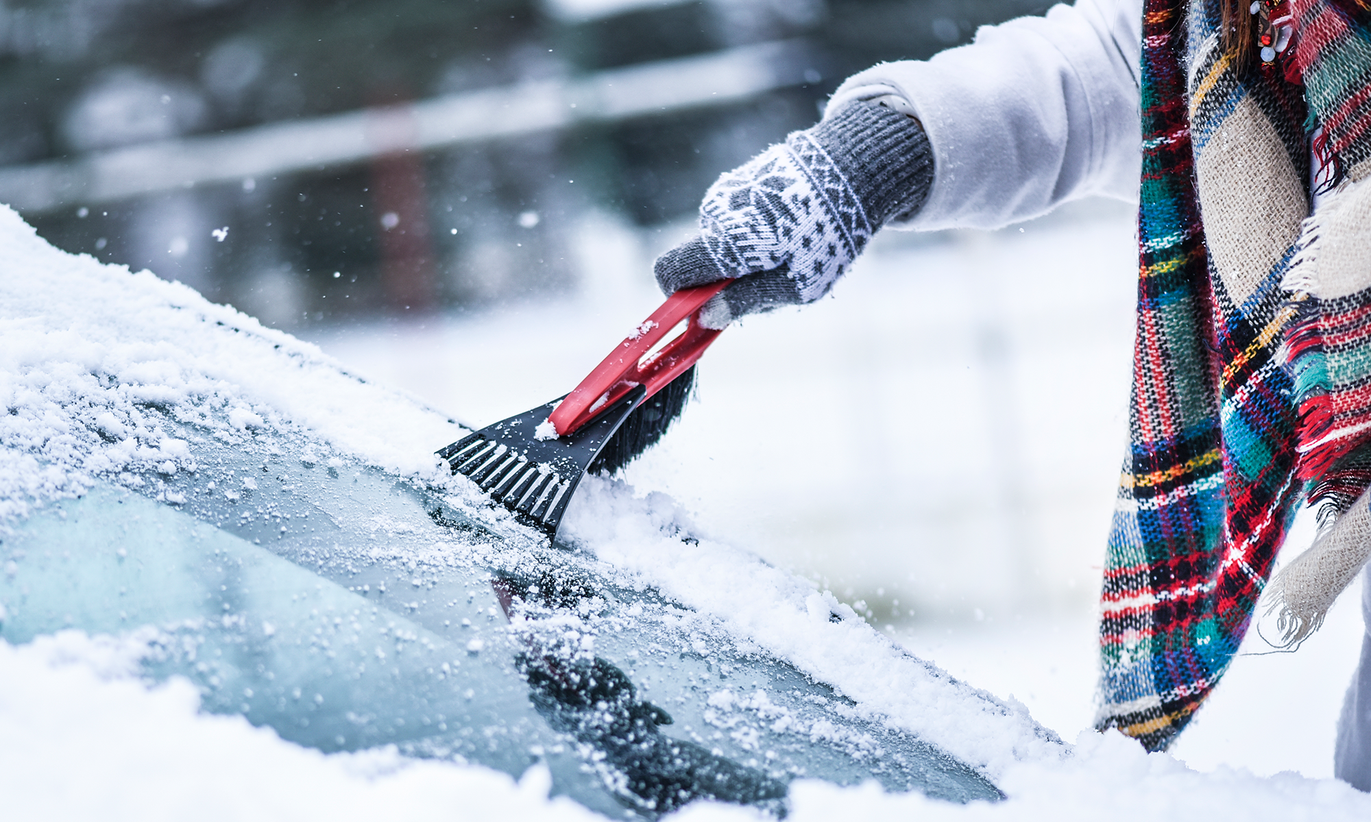 Un primer plano de una persona raspando el parabrisas de su automóvil con un raspador de hielo mientras viste ropa, una bufanda y una chaqueta de invierno blanca.
