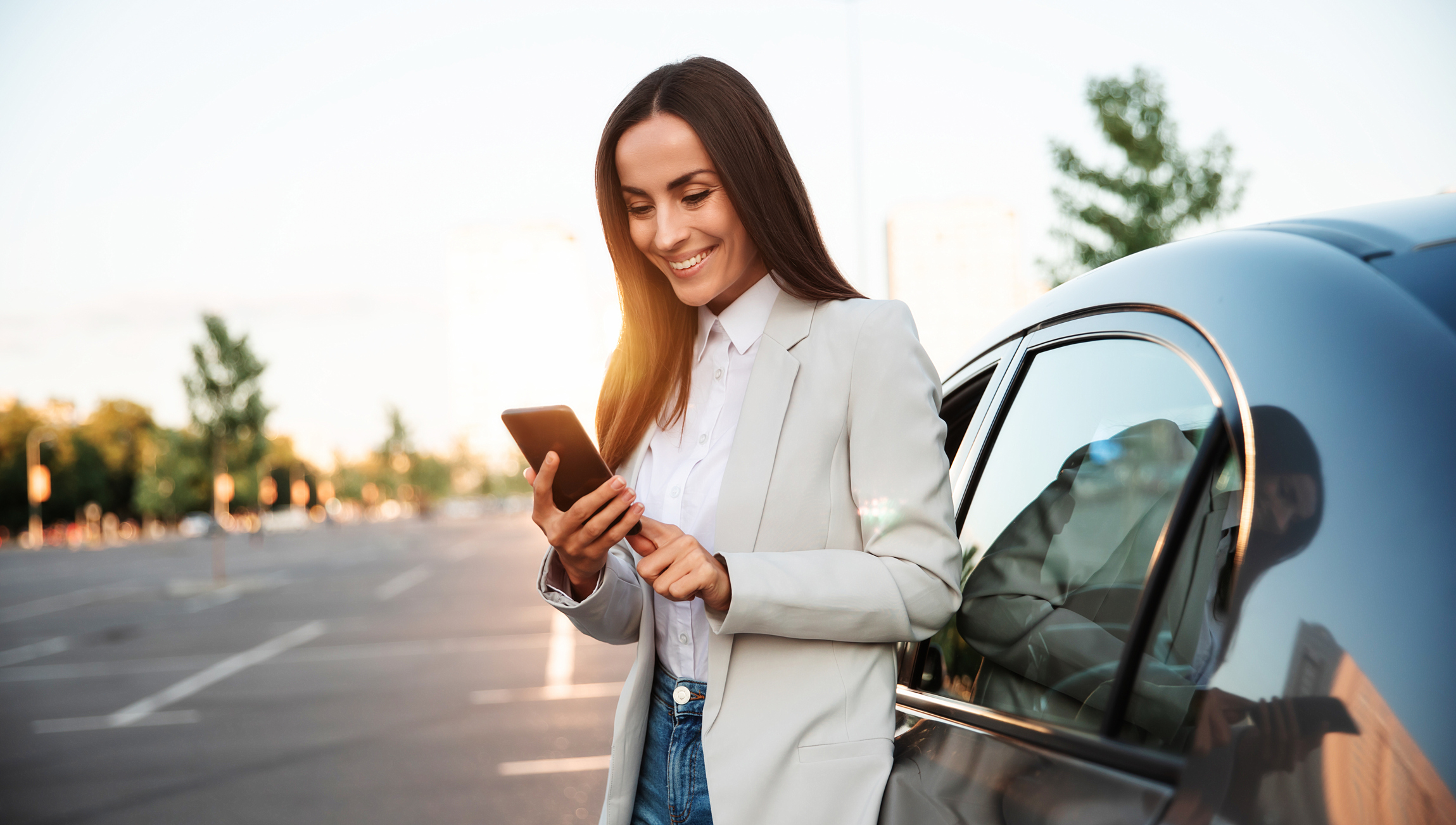 Successful smiling attractive woman in formal smart wear is using her smart phone while standing near modern car outdoors