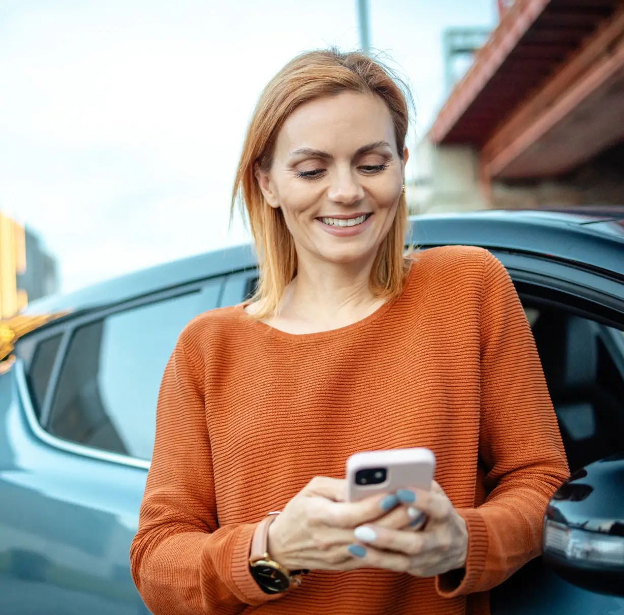 Woman in orange shirt smiling at her phone while standing next to her SUV